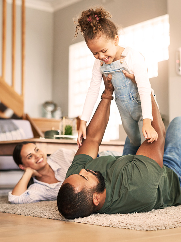 Eine junge Familie liegt auf dem Teppichboden. Der Vater hebt seine Tochter mit beiden Händen in die Luft. Alle lachen.